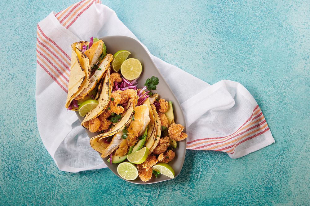 Shrimp tacos served with limes on a long gray plate on top of a white towel and a vibrant blue background.