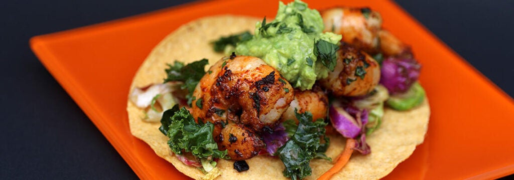 Shrimp tacos with guac, kale, and many other toppings, served on a crunchy shell on a square orange plate.