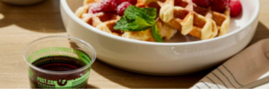 Waffles in a bowl with raspberries on them and a small compostable cup of syrup next to it.