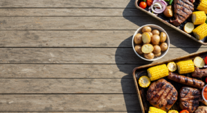 A grilled feast featuring steak, sausages, corn on the cob, and potatoes is laid out on a rustic wooden table.