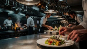 Scène de cuisine professionnelle occupée montrant quatre chefs en train de travailler, l’un d’eux préparant un plat de poisson et de légumes au premier plan