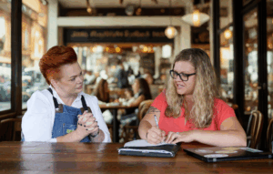 Two women sit at a wooden table in a brightly lit cafe, with one wearing denim overalls and listening intently while the other, wearing a red shirt and glasses, writes in a notebook.
