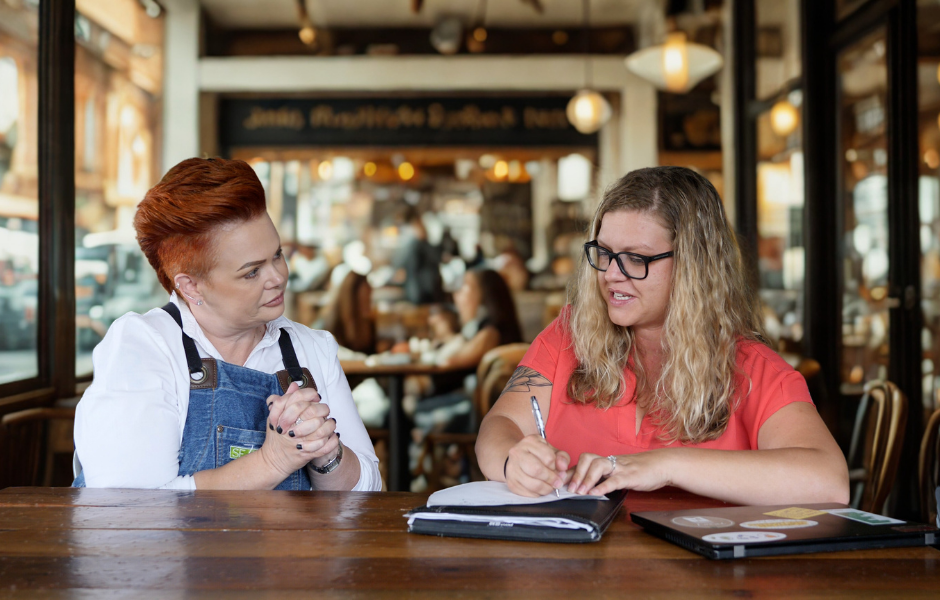 Two women sit at a wooden table in a brightly lit cafe, with one wearing denim overalls and listening intently while the other, wearing a red shirt and glasses, writes in a notebook.
