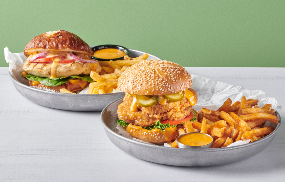 Two chicken sandwiches served on metal trays with seasoned fries and dipping sauce, featuring one grilled chicken sandwich on a pretzel bun and one fried chicken sandwich on a sesame seed bun.
