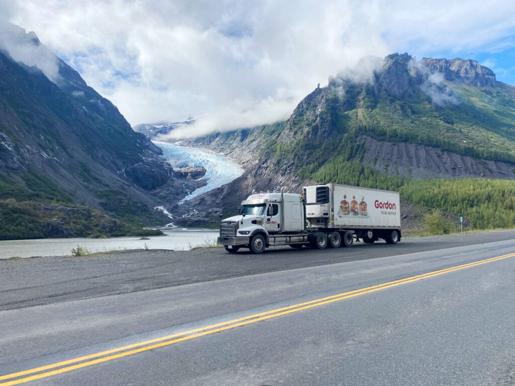 A Gordon Food Service semi-truck is parked on the shoulder of a paved highway, set against a dramatic backdrop of a turquoise glacier flowing between mist-cover A Gordon Food Service semi-truck is parked on the shoulder of a paved highway, set against a dramatic backdrop of a turquoise glacier flowing between mist-covered mountains.ed mountains.