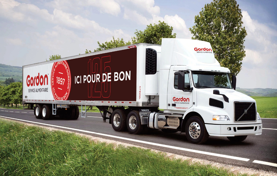 Un semi-remorque blanc de Gordon Food Service, dont la remorque est ornée d'un hamburger, est garé sur le quai de chargement d'un centre de distribution dont la ligne de toit rouge est proéminente.