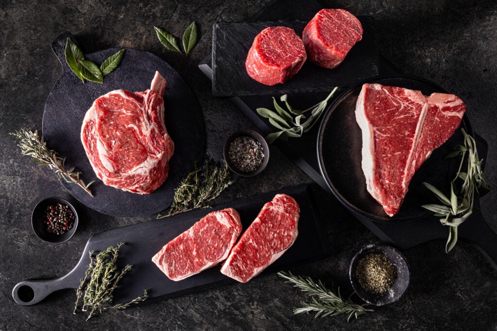 Une sélection de steaks de bœuf marbrés, épais et non affinés, comprenant notamment des côtes de bœuf, des filets mignons, des T-bone et des faux-filets, est disposée avec art sur des planches à découper et des plateaux sombres, accompagnée d'herbes fraîches et de petits bols d'épices.