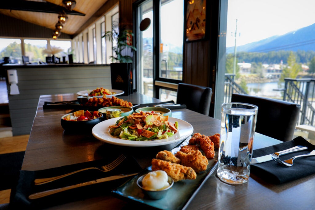 A dining table featuring crispy chicken wings, fresh salad, appetizers, and water next to large scenic resort windows.