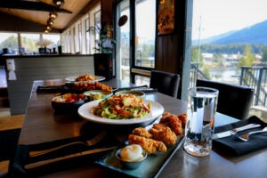 A dining table featuring crispy chicken wings, fresh salad, appetizers, and water next to large scenic resort windows.